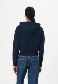 Felpa blu navy corta con cappuccio a coulisse e polsini a costine. Abbinata a jeans blu a vita alta con tasche posteriori e texture scolorita.