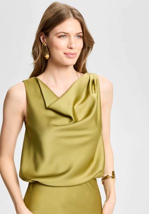 Femme portant un top en satin vert olive drapé sans manches avec de grandes boucles d'oreilles dorées en forme d'anneaux et un bracelet manchette doré.