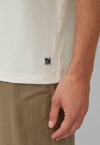 Kremowa bawełniana koszulka z małą granatową metką z napisem "s.O." na dołku, w zestawie z teksturowanymi brązowymi spodniami.