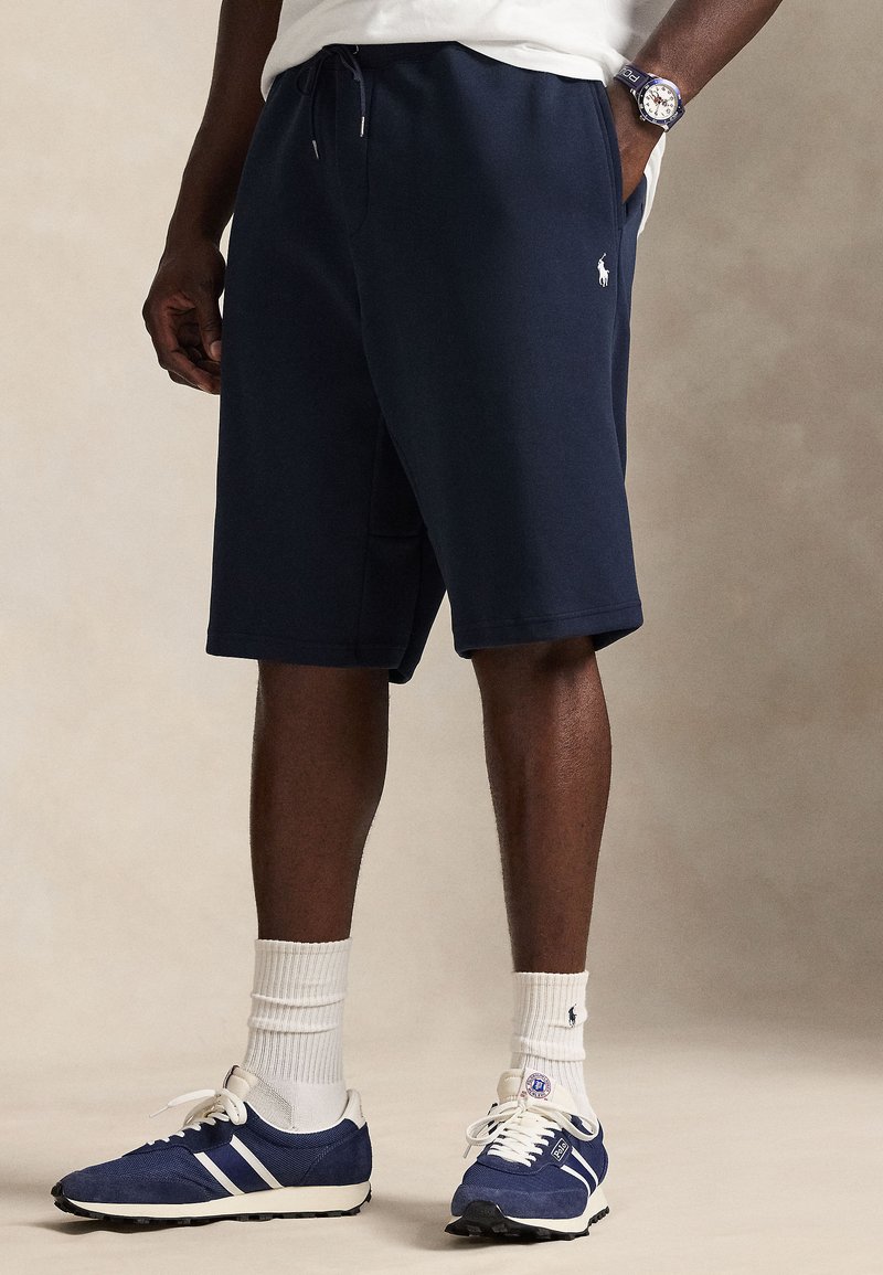 Σκούρο μπλε αθλητικό σορτς από μαλ ακό βαμβάκι με πλαϊνές τσέπες και ελαστική ζώνη μέσης, συνδυασμένο με σκούρα μπλε και λευκά παπούτσια και λευκές κάλτσες.