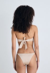 Beige bikini, hátra megkötős toppal és minimális takarást biztosító alsó résszel. Simaság, egyszerű dizájn és állítható pántok hátul.