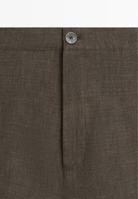 Close-up af linning på mørkebrune linnedbukser med en enkelt grå knap og synlige sømme omkring knaphullet og gylpen.