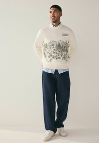 Cream sweatshirt with colorful, detailed mountain scene; dark blue trousers; white sneakers; layered with light blue shirt underneath.