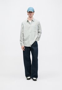 Ungdomsmann iført lys blå baseballcap, løs rutete skjorte med knapper, vide mørke jeans og svart-hvite sko, står foran hvit bakgrunn.