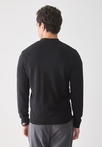 Mann, der von hinten zu sehen ist, trägt einen schlichten schwarzen Langarmpullover und graue Hosen vor einem hellgrauen Hintergrund.