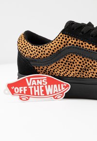 Baskets Vans tachetées noir et fauve avec bande noire, semelle noire et étiquette rouge Vans « Off The Wall » en forme de planche de skateboard sur fond blanc.