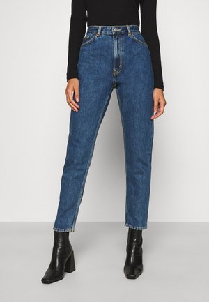 Kvinde iført højttaljede blå jeans, sort langærmet top og sorte ankelstøvler med hæl, stående mod en ensfarvet baggrund.