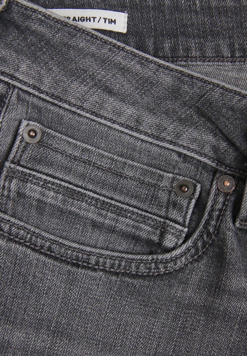 Jeans de mezclilla gris con un bolsillo frontal con un botón de metal, costuras contrastantes y una etiqueta que indica la marca y el estilo.