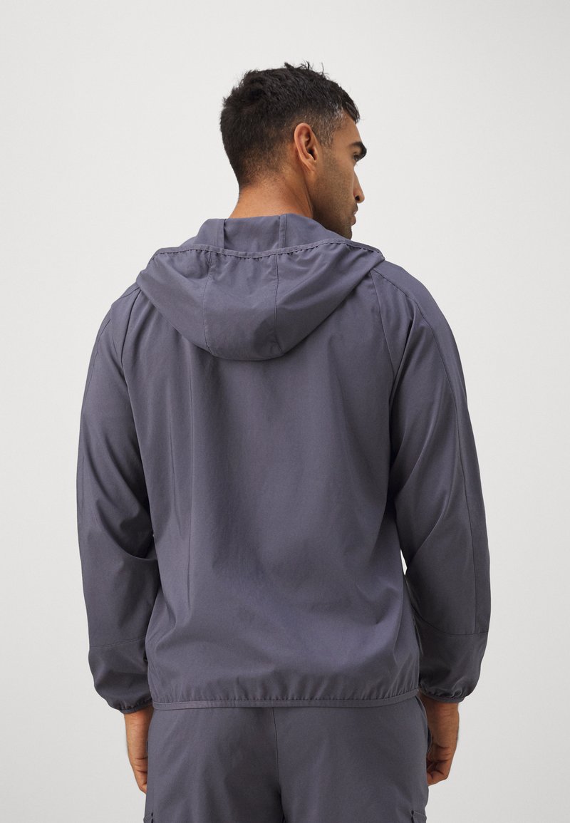Grå letvægtsjakke med hætte, elastiske manchetter og løs pasform. Glat tekstur, minimalistisk design, skræddersyet til komfort og bevægelse.