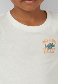 Weißes Baumwoll-T-Shirt mit rundem Ausschnitt und kurzen Ärmeln, mit einem bunten Grafik eines Dinosauriers und dem Text "WILD CLAWS VALLEY."