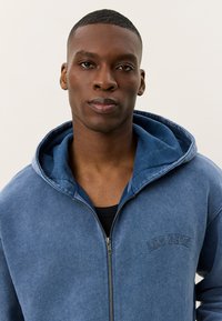 Blauwe rits hoodie gemaakt van zachte stof, met een gevoerde capuchon, geribde manchetten en een subtiele borstgrafiek. Glad textuur met een ontspannen pasvorm.