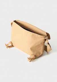 Beige myk stoffveske med glidelås på toppen, flat rektangulær form og justerbare stropper på begge sider. Minimalistisk design uten synlige logoer.