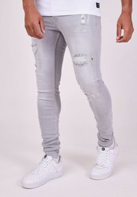 Jeans skinny gris claro con detalles desgastados y bordes deshilachados, combinados con zapatillas blancas. Enfócate en la textura y el ajuste.