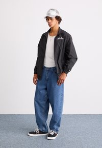 Čierna ľahká bunda s elastickými manžetami, biela tričko, voľné modré džínsy a čierne tenisky s bielymi šnúrkami a červenými akcentmi.