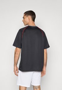 T-shirt de sport gris foncé à manches courtes, avec des accents rouges le long des épaules et un col blanc. Porté avec un short blanc.