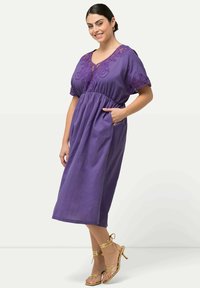 Violetti puuvillamekko lyhyillä hihoilla, brodeerattu kaula-aukko ja sivutaskut. Ominaisuuksiin kuuluu rento malli ja se ulottuu polven alapuolelle. Kultaisten nilkkaketjujen sandaalit.
