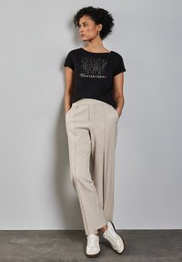 Žena s krátkými kudrnatými vlasy, oblečená v černém tričku s nápisem "Spokojenost" a béžových kalhotách, stojí u světle šedé zdi.