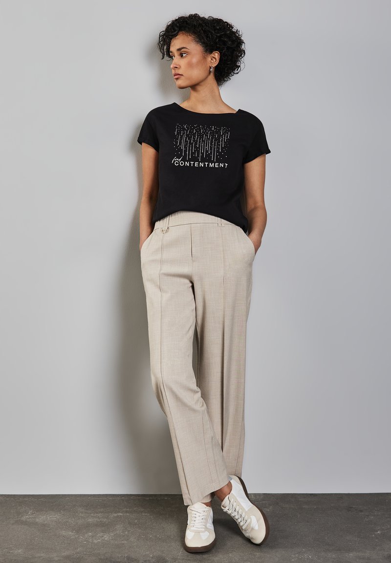 Žena s krátkými kudrnatými vlasy, oblečená v černém tričku s nápisem "Spokojenost" a béžových kalhotách, stojí u světle šedé zdi.