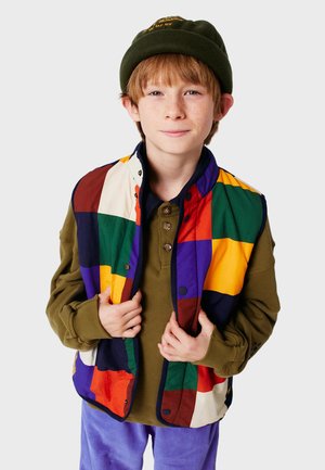Gilet coloré en patchwork avec des carrés rouges, verts, jaunes, violets et blancs. Porté sur une chemise verte, avec trois boutons visibles.