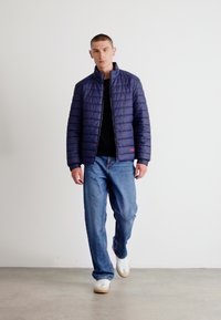 Marineblå quilted jakke med kort oppreist krage, svart topp under, kombinert med løse blå jeans og hvite sneakers. Minimalistisk design.
