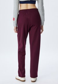 Pantalones de chándal burdeos con cintura elástica y bolsillos laterales, que cuentan con una textura suave y un ajuste tapered. Se llevan zapatillas blancas.