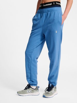 Osoba ubrana w niebieskie spodnie dresowe i szare buty sportowe, stojąca z jedną ręką w kieszeni na jednolitym białym tle.