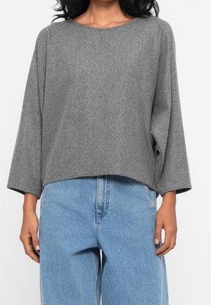 Žena nosící volné šedé tričko s dlouhým rukávem a širokými rukávy v kombinaci s světle modrými džíny s vysokým pasem.