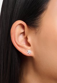 Weiße sternförmige Ohrringe mit einem kleinen zentralen Edelstein, vor einer Nahaufnahme eines Ohrs und einem Hintergrund aus glattem dunklem Haar.