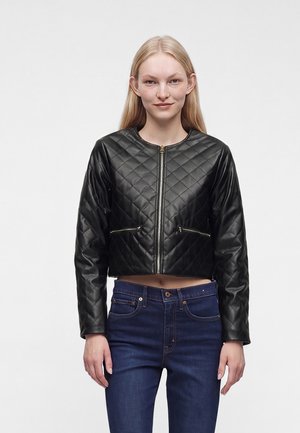 Jeune femme aux cheveux blonds raides porte une veste en cuir noire matelassée, courte, et un jean taille haute bleu foncé.
