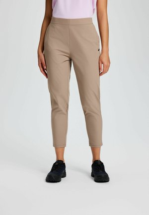 Frau trägt beige kurze Hosen und schwarze Wanderschuhe, steht vor einem schlichten hellen Hintergrund.