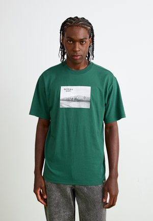 Tricou din bumbac verde, cu mâneci scurte, având un imprimeu grafic rectangular alb-negru și textul "DICKIES 1922" la centru.