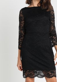 Svart spetsklänning med trekvartslånga ärmar, blommönster, vågig kant och en figurnära siluett. Texturerad tyg ger djup.