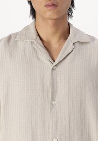 Giovane che indossa una camicia button-up beige chiaro con texture e colletto a revers, mostrato dalla nuca fino al mezzo torso su uno sfondo bianco.