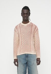 Pull en maille avec un motif texturé crème et rouge rouille. Comprend un col rond et des poignets côtelés, associé à un jean bleu clair.