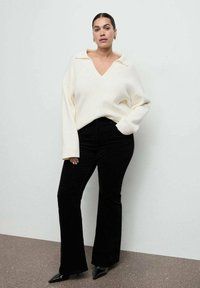 Biały prążkowany sweter z dekoltem w serek i szerokim kołnierzem, zestawiony z czarnymi spodniami dzwonami. Modelka ma na sobie czarne szpilki w szpic oraz kolczyki koła.