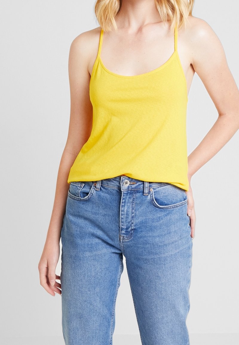 Gul ärmlös topp i lätt tyg med tunna axelremmar, i kombination med högmidjade blå jeans. Toppen har en avslappnad passform.