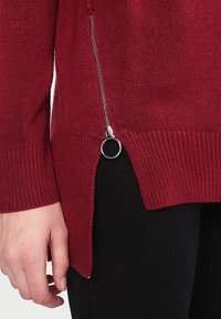Bordeauxfarbener Pullover mit geripptem Muster, ausgestattet mit einem seitlichen Reißverschluss und einem gespaltenen Saum, kombiniert mit schwarzem Stoff.