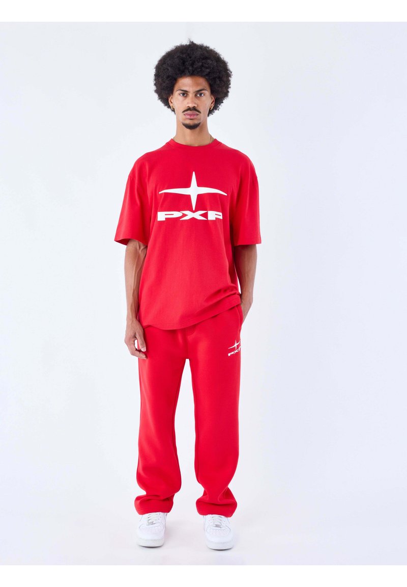 Homme avec coiffure afro portant un t-shirt rouge avec logo PXP et un pantalon rouge assorti avec des baskets blanches, debout sur fond blanc.
