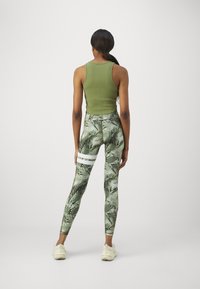 Oliwkowy top na ramiączkach i dopasowane legginsy w zielony liściasty wzór. Legginsy mają dwa białe poziome paski na górnej części nóg.