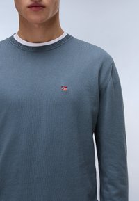 Lichtblauw gebreid sweater met een ronde halslijn en ribbelmanchetten. Bevat een klein rood en wit vlagembleem op de borst. Zachte textuur.