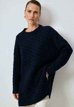 Donna che indossa un maglione blu navy a trecce con scollatura rotonda, orecchini a cerchio dorati e pantaloni bianchi, in piedi davanti a un muro semplice.