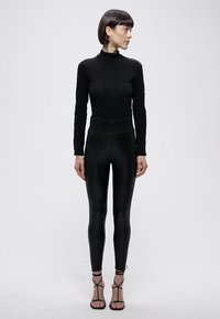 Crna majica dugih rukava s visokim ovratnikom i detaljem od čipke, uparena s crnim visokim tajicama i sandalama s remenčićima. Glatka tekstura.