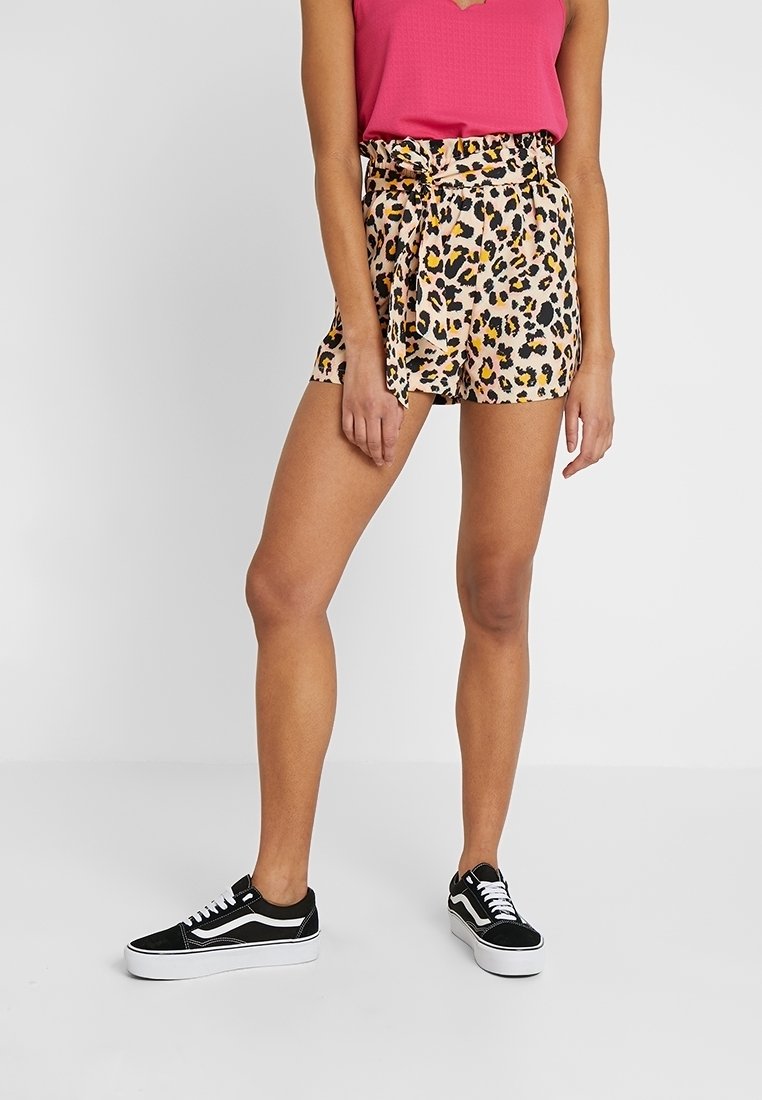 Femme portant un débardeur rose, un short taille haute à imprimé léopard avec ceinture nouée, et des baskets basses noires à semelles blanches.