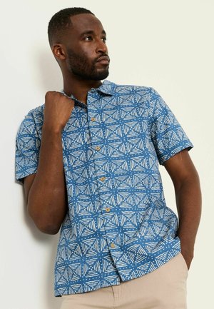 Mann trägt ein blau gemustertes kurzärmeliges Hemd mit Knöpfen und beige Hose, posiert mit einer Hand am Kragen und der anderen in der Tasche.