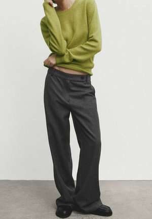 Personne portant un pull vert citron en tissu duveteux et un pantalon ample gris foncé, debout sur un sol gris devant un mur blanc.