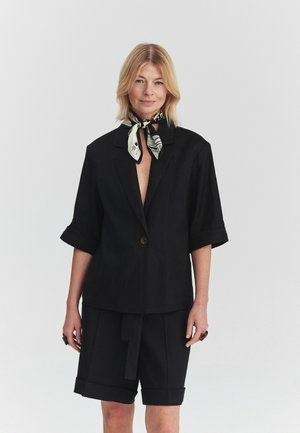Lyshåret kvinde iført sort blazer med korte ærmer og matchende knælange shorts, accessoriseret med et sort-hvidt mønstret halstørklæde.