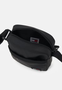 Borsa a tracolla nera con chiusura superiore con zip, superficie liscia, tracolla regolabile e un patch frontale con dettagli rossi, bianchi e blu.