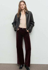 Chaqueta de cuero negra, blusa beige abotonada, pantalones de pana burdeos de pierna ancha y botas de tacón negras. Texturas suaves, colores contrastantes.