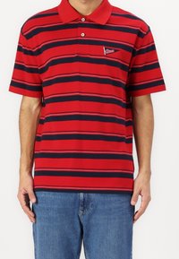 Polo à rayures rouges avec rayures horizontales bleu marine, col avec deux boutons, et un petit logo sur la poitrine gauche. Assorti avec un jean bleu.
