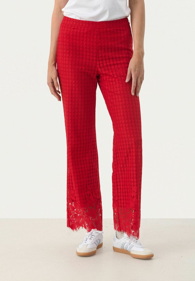 Femme portant un pantalon rouge texturé avec des ourlets en dentelle et des baskets blanches, debout devant un fond clair uni.